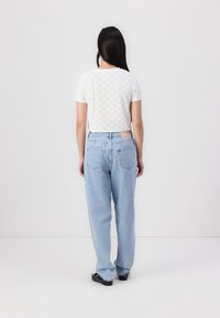 Vit kortärmad skjorta med broderat mönster, tillsammans med ljusblå, löst sittande jeans och svarta skor, sedd från baksidan.