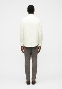 Camisa de manga comprida de cor clara com corte reto, combinada com calças castanhas texturizadas. A camisa tem punhos com botões; sem padrões visíveis.