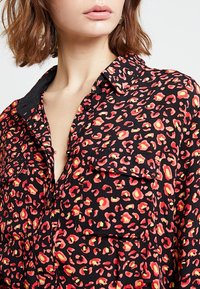 Chemise noire avec imprimé léopard rouge et jaune, dotée d'un col, d'une fermeture à boutons sur le devant, de deux poches poitrine et d'une texture douce.