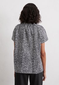 Personne aux cheveux bouclés et foncés portant un chemisier à manches courtes noir à taches blanches et un pantalon noir, debout devant un mur blanc uni.