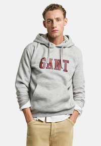 Felpa grigia con cappuccio, caratterizzata da un grande logo rosso "GANT" sul davanti e una tasca a marsupio. Una camicia bianca spunta da sotto.
