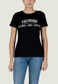 T-shirt en coton noir avec un graphique blanc "GUESS AMERICAN TRADITION 1981" agrémenté d'accents argentés et une coupe ajustée. Manches courtes.
