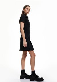 Schwarzes kurzärmeliges Kleid mit geknöpftem Vorderteil, plissiertem Rock und tailliertem Oberteil. Getragen mit glänzenden schwarzen Plateauschuhen und schwarzen Socken.
