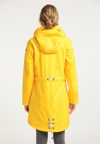 Kobieta o blond włosach związanych w kok, ubrana w żółty płaszcz z kapturem w białe wzory kotwicy i regulowanymi białymi troczkami, zwrócona tyłem.