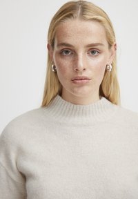 Maglione lavorato a maglia in una tonalità beige chiaro con colletto alto a coste. Tessuto morbido con vestibilità comoda. Il modello indossa orecchini a cerchio argentati.