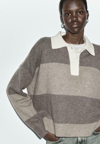 Pull avec de larges rayures horizontales en marron clair et beige, doté d'un col blanc contrastant et d'une patte de boutonnage, fabriqué en matériau tricoté doux.