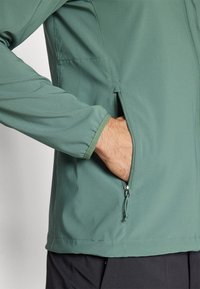 Ręka wsunięta do zasuwanej bocznej kieszeni zielonej kurtki, widoczny elastyczny mankiet rękawa i część czarnych spodni.