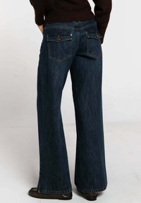 Jean denim bleu foncé à jambes larges avec deux poches arrière et rivets en cuivre. Texture lisse et coupe droite de la taille jusqu'aux chevilles.