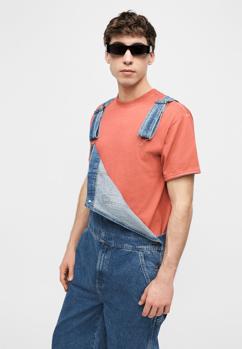 Giovane uomo che indossa occhiali da sole neri, una maglietta corallo e una salopette di denim blu con una spallina slacciata su uno sfondo grigio uniforme.