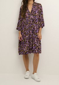 Vestido con patrón floral en púrpura, rosa y verde sobre fondo marrón. Cuello en V, mangas acampanadas tres cuartos, falda de volantes, combinado con zapatillas blancas.