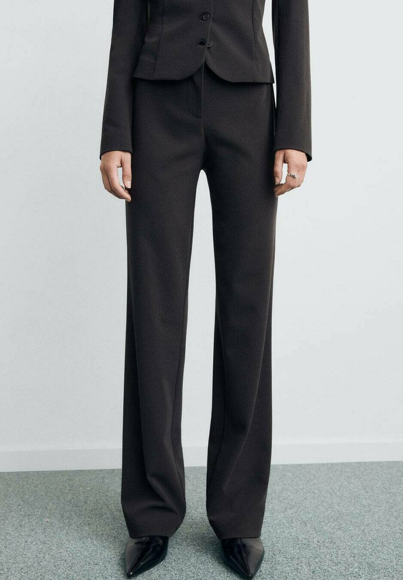 Pantaloni neri su misura con una vestibilità a gamba larga, texture liscia e vita alta. Indossati con tacchi neri a punta e una giacca aderente.