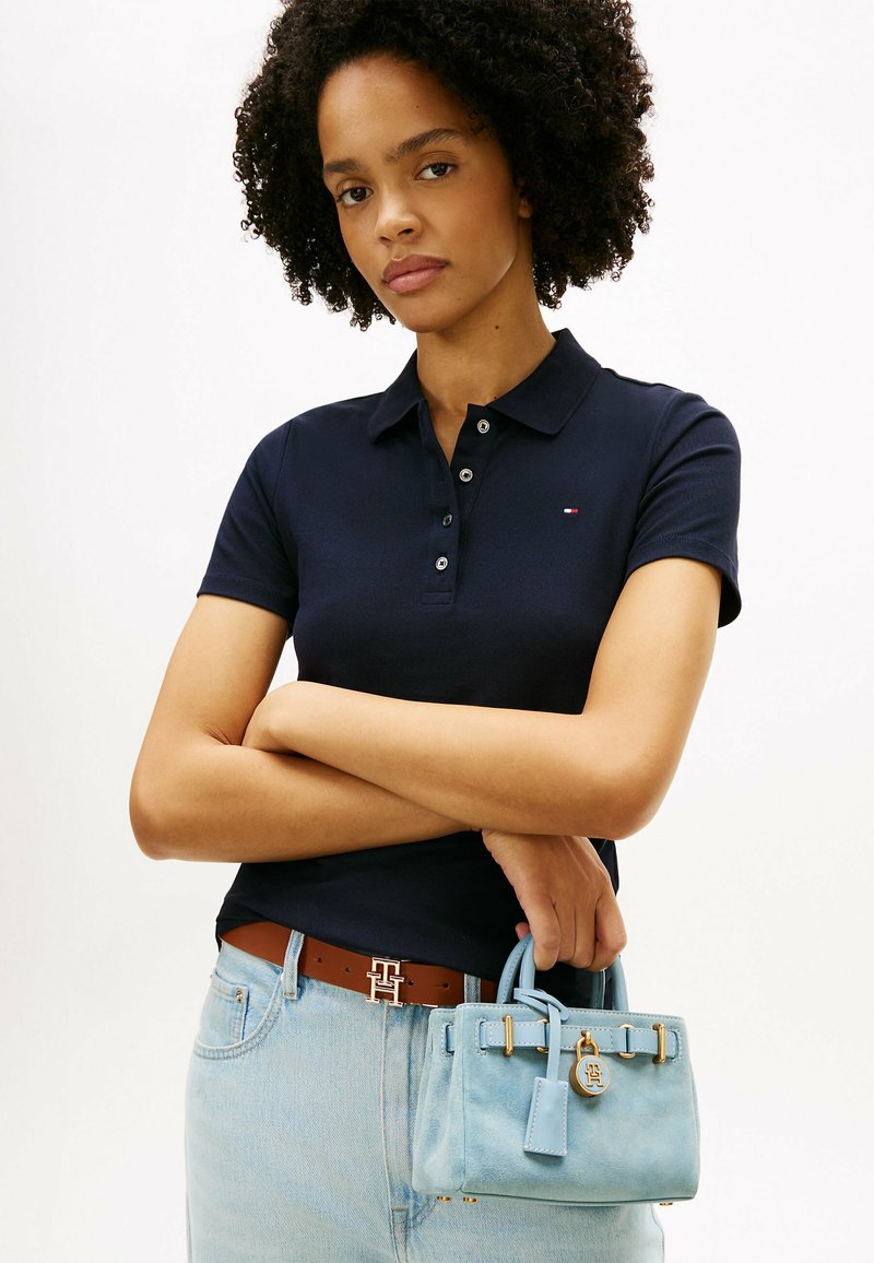 Mujer con cabello rizado que lleva una camiseta polo azul marino y jeans azul claro sostiene un pequeño bolso azul claro con detalles dorados.