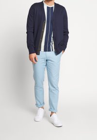 Muž v námořnickém cardigan, pruhovaném námořnickém a bílém tričku, světle modrých zkrácených kalhotách a bílých teniskách na bílém pozadí.