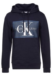 Tmavě modrá mikina s kapucí vyrobená z bavlněné směsi, která má na přední straně velký modro-bílý grafický potisk s nápisem "CK CALVIN KLEIN JEANS" a kapsu typu klokanka.