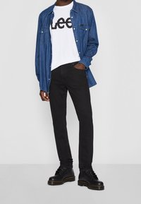 Denim shirt with snap buttons, blue color, worn over a white t-shirt with black logo. Black slim-fit jeans and black lace-up boots.