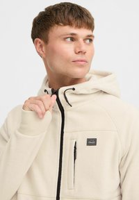 Beige zip-up fleece hættejakke med en sort lynlås, snøre i huen og frontlomme. Har et brandlogo på øverste venstre side.