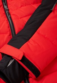 Manche de veste matelassée rouge avec poignet intérieur noir et sangle Velcro ajustable près du poignet et détail de fermeture éclair noire sur tissu rouge.