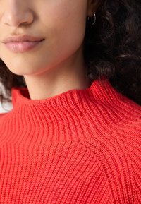 Strickpullover in leuchtendem Rot mit einem hohen ribbeligen Kragen und strukturiertem Muster. Nahaufnahme, die die Details des Stoffes hervorhebt.