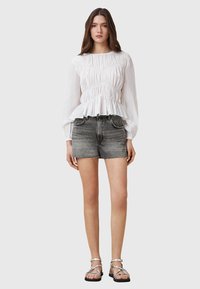 Blouse blanche à manches longues avec texture plissée et ourlet en peplum, associée à un short en denim noir et des sandales argentées à lanières.