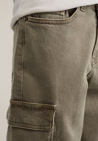 Maslinasto zelene cargo kratke hlače od teksturiranog materijala. Sadrže bočne džepove s patenticama i ukrasne šavove. Nose se s bijelom košuljom.