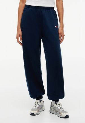 Osoba nosząca granatowe spodnie dresowe z elastycznymi ściągaczami i logo Tommy Hilfiger na lewym udzie, w połączeniu z białymi i szarymi sneakersami.