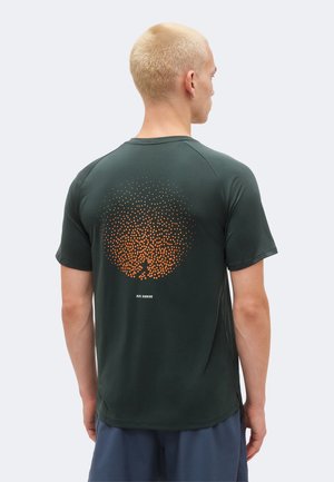 Rövid szőke hajú férfi, sötétzöld rövid ujjú pólóban, amelyen narancssárga pöttyös kör és a „Nike Running” felirat található a hátulján.