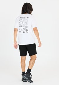 Vit t-shirt med illustrerade grafiker på baksidan, kombinerad med svarta shorts och svarta vandringskor. Modell sedd bakifrån.