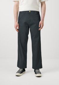 Pantalons larges gris foncé, avec une texture lisse, présentant une fermeture à bouton et des passants de ceinture, associés à des baskets en toile noires.