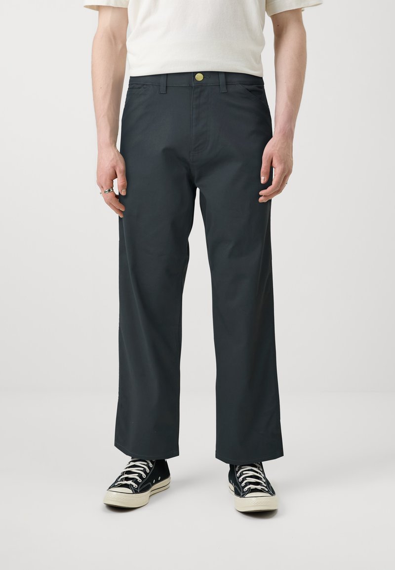 Pantalons larges gris foncé, avec une texture lisse, présentant une fermeture à bouton et des passants de ceinture, associés à des baskets en toile noires.