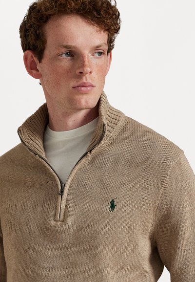 Beige lynlåstrøje med ribbet krave, struktureret strikstof og et lille grønt logo på venstre bryst. Lagt over en lys t-shirt.