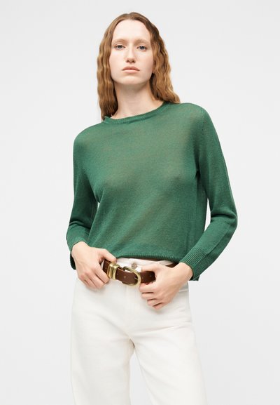 Femme aux cheveux ondulés portant un pull vert en maille et un pantalon blanc, ajustant une ceinture marron avec une grande boucle dorée.