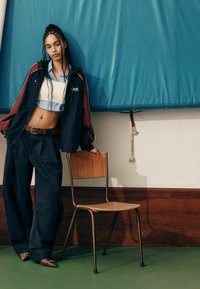 Tenue streetwear à la mode : veste marine et bordeaux, crop top bleu clair, pantalon ample noir, et chaussures texturées marron. Chaise en bois à proximité.