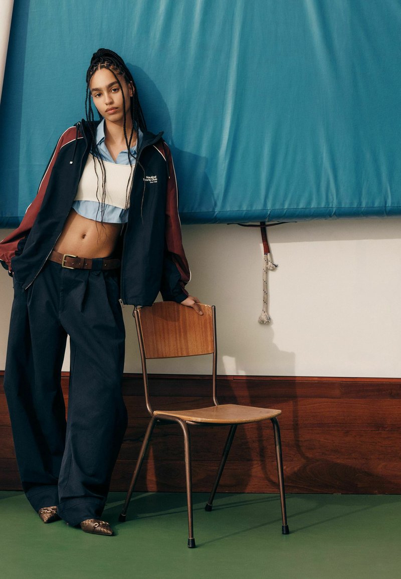Tenue streetwear à la mode : veste marine et bordeaux, crop top bleu clair, pantalon ample noir, et chaussures texturées marron. Chaise en bois à proximité.
