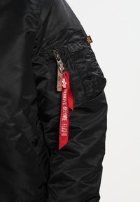 Veste bomber noire à finition brillante, avec une poche zippée sur la manche et une étiquette rouge "REMOVE BEFORE FLIGHT" attachée.