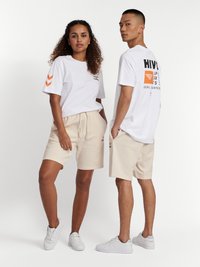 Vita t-shirts med grafiska tryck och orange detaljer, kombinerade med beigea shorts. Båda bär vita sneakers, vilket visar upp en avslappnad och bekväm stil.