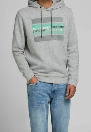 Mężczyzna w jasnoszarej bluzie z kapturem z napisem „JACK & JONES 1990 CORE” i jasnoniebieskich dżinsach, jedną ręką w kieszeni, neutralne tło.