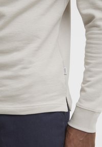 Long-sleeve light beige shirt made of soft textile, featuring a side slit and ribbed cuffs, with a small brand tag near the seam.