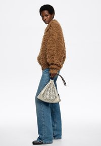 Chaqueta marrón esponjosa, pantalones vaqueros de pierna ancha azules y una bolsa de cuerda crema con un patrón floral negro. La modelo posa de perfil.