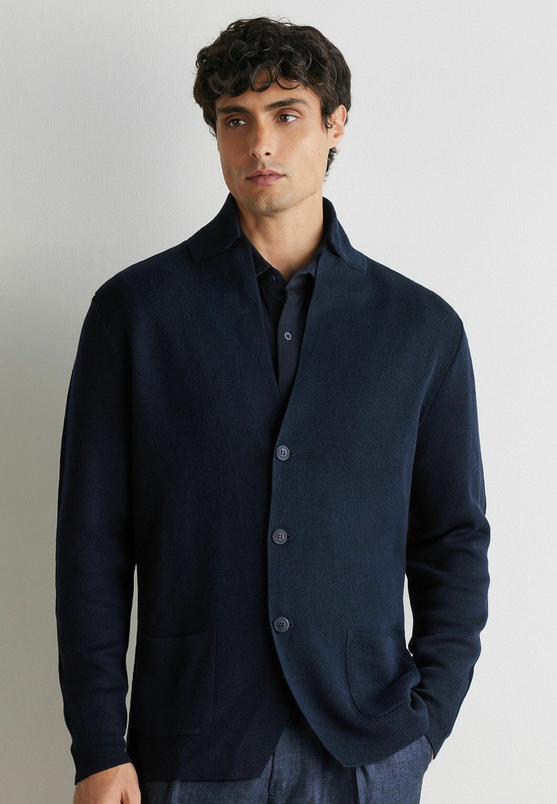 Młody mężczyzna z kręconymi włosami, ubrany w ciemnogranatowy, zapinany na guziki sweter na tle jasnego, jednolitego tła.