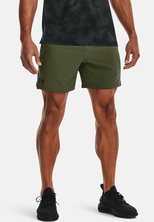 Sportliche Person in olivgrünen Shorts, schwarzen Sportschuhen und einem schwarzen Shirt, steht mit gespreizten Beinen vor weißem Hintergrund.