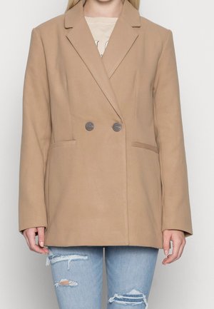 Kobieta w beżowej dwurzędowej marynarce na białej koszuli i podartych jasnoniebieskich dżinsach, stojąca na tle jednolitego tła.