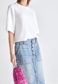 Un haut blanc à manches courtes côtelé associé à une jupe en denim bleu clair ornée d'un bouton et de poches latérales, le tout complété par un sac à main à franges roses.