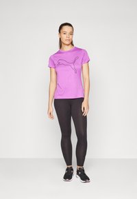 Lila kortärmad t-shirt med en svart kattlogotyp, parat med svarta leggings och svarta sneakers. Slät textur och atletisk passform.