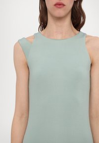 Haut sans manches vert clair avec une texture lisse, présentant une coupe d'épaule asymétrique unique et un col rond.