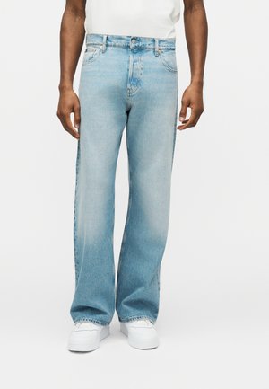 Mand iført lyseblå vide jeans og hvide sneakers, stående mod en ensfarvet hvid baggrund.