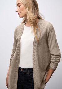 Cárdigan de punto beige con un frente abierto acanalado, mangas largas y una textura suave, combinado con una blusa blanca y jeans oscuros.
