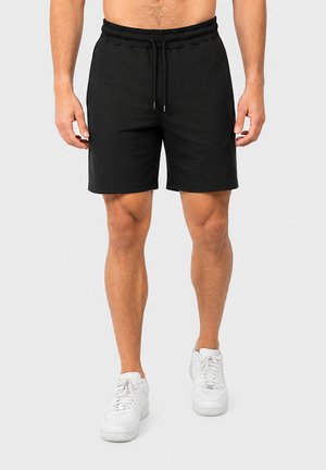 Homme portant un short noir uni avec cordon et des baskets blanches, debout devant un fond gris, torse et jambes visibles.