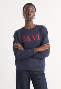 Naminis mėlynas džemperis, pagamintas iš medvilnės ir sintetinių pluoštų, su raudonu išsiuvinėtu "GANT" logotipu per krūtinę, turintis banguotą apykaklę ir rankogalius.