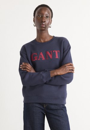 Hanorac de culoare bleu marin dintr-un amestec de bumbac, având logo-ul roșu brodat "GANT" pe piept, cu guler și manșete ribbate.
