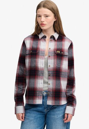 Jonge vrouw die een rood, zwart en wit geruit overhemd met knopen draagt over een lichtgrijze top en blauwe jeans, staande tegen een effen achtergrond.